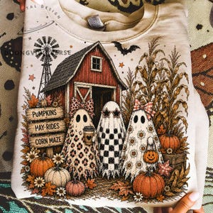 Könnte beinhalten: Ein beigefarbenes Sweatshirt mit Rundhalsausschnitt und einem Grafikdruck einer roten Scheune, einer Windmühle und drei Geistern. Die Geister tragen Accessoires wie eine Leoparden-Schleife, sternförmige Sonnenbrillen und eine Blumen-Schleife. Text: PUMPKINS, HAY-RIDES, CORN MAZE.