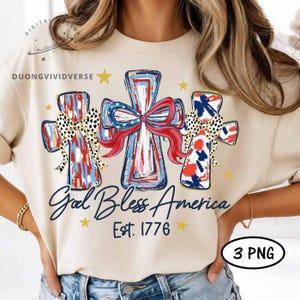 May include: A beige t-shirt featuring a graphic of three crosses in red, white, and blue with leopard print bows and a red ribbon. The text reads God Bless America Est. 1776, accompanied by yellow stars. The design includes the text DUONGVIVIDVERSE.