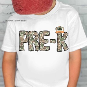 May include: A white t-shirt featuring the text PRE-K in a camouflage pattern with black stitched outlines. A small graphic of a baseball cap with the text BACK TO SCHOOL is positioned above the letter K. The design is centered on the chest.