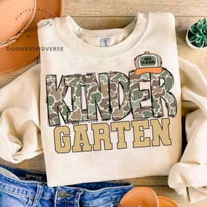 May include: A cream-colored crewneck sweatshirt featuring the word KINDER in a green and brown camouflage pattern with black stitched outlines, and the word GARTEN in solid tan letters below. A small orange and tan baseball cap icon says BACK TO SCHOOL.