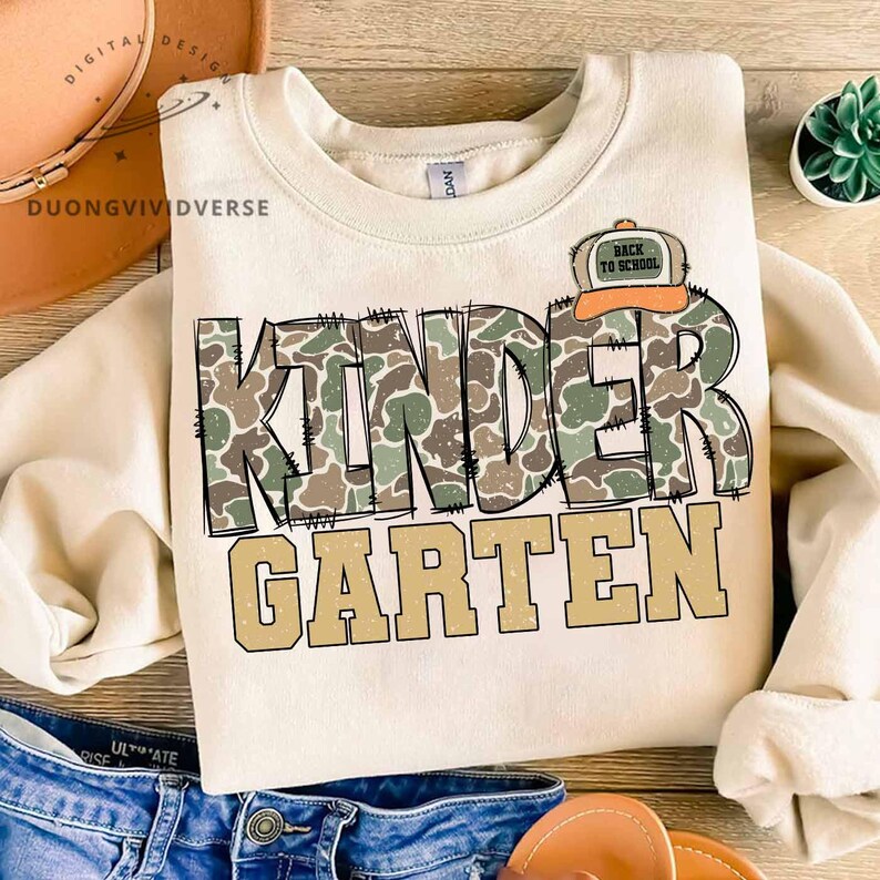 May include: A cream-colored crewneck sweatshirt featuring the word KINDER in a green and brown camouflage pattern with black stitched outlines, and the word GARTEN in solid tan letters below. A small orange and tan baseball cap icon says BACK TO SCHOOL.