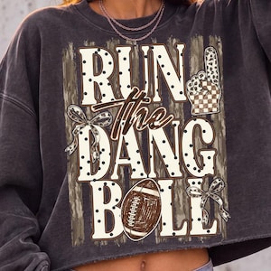 May include: A charcoal gray cropped sweatshirt featuring a graphic print with the text RUN THE DANG BALL. The design includes a brown football, a checkered foam finger, and two leopard print bows against a distressed tan background.