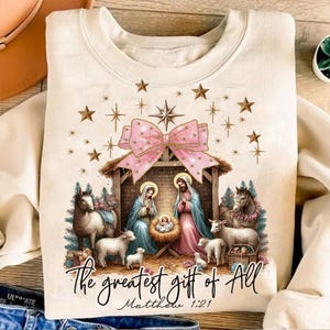 May include: A beige sweatshirt with a Christmas nativity scene design. The scene features Mary, Joseph, and baby Jesus in a manger. There are two sheep and a donkey in the scene. The text "The greatest gift of all Matthew 1:21" is printed below the scene.