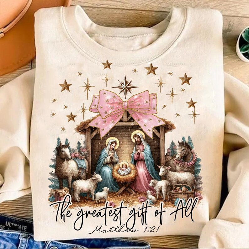 May include: A beige sweatshirt with a Christmas nativity scene design. The scene features Mary, Joseph, and baby Jesus in a manger. There are two sheep and a donkey in the scene. The text "The greatest gift of all Matthew 1:21" is printed below the scene.