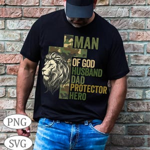 Może przedstawiać: Czarny t-shirt z grafiką lwa i napisem "MAN OF GOD HUSBAND DAD PROTECTOR HERO" w kolorze zielonym i moro. Koszulka jest noszona przez osobę w czapce i niebieskich dżinsach.