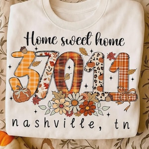 May include: A white crewneck sweatshirt featuring the text Home sweet home at the top and nashville, tn at the bottom. The center displays the number 37011 in large, patterned digits decorated with plaid, leopard print, pumpkins, and autumn leaves.