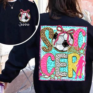 Op de afbeelding: Een marineblauw sweatshirt met een voetbalafbeelding met een roze strik en het woord "Jessie" op de voorkant. De achterkant toont het woord "SOCCER" met een voetbal, een silhouet van een speler en steraccenten.
