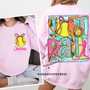 May include: Pink sweatshirt with a colourful graphic design featuring a softball, a bat, and the word "Softball". The graphic also includes stars, animal print, and a silhouette of a player. The sweatshirt has the name "Jessie" printed on the front.