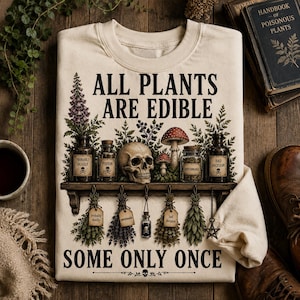 May include: Cream-colored sweatshirt with the text "ALL PLANTS ARE EDIBLE SOME ONLY ONCE". The design includes a shelf with a skull, mushrooms, and jars labeled "Poison" and "Bad Decision". Hanging below are bundles of herbs.