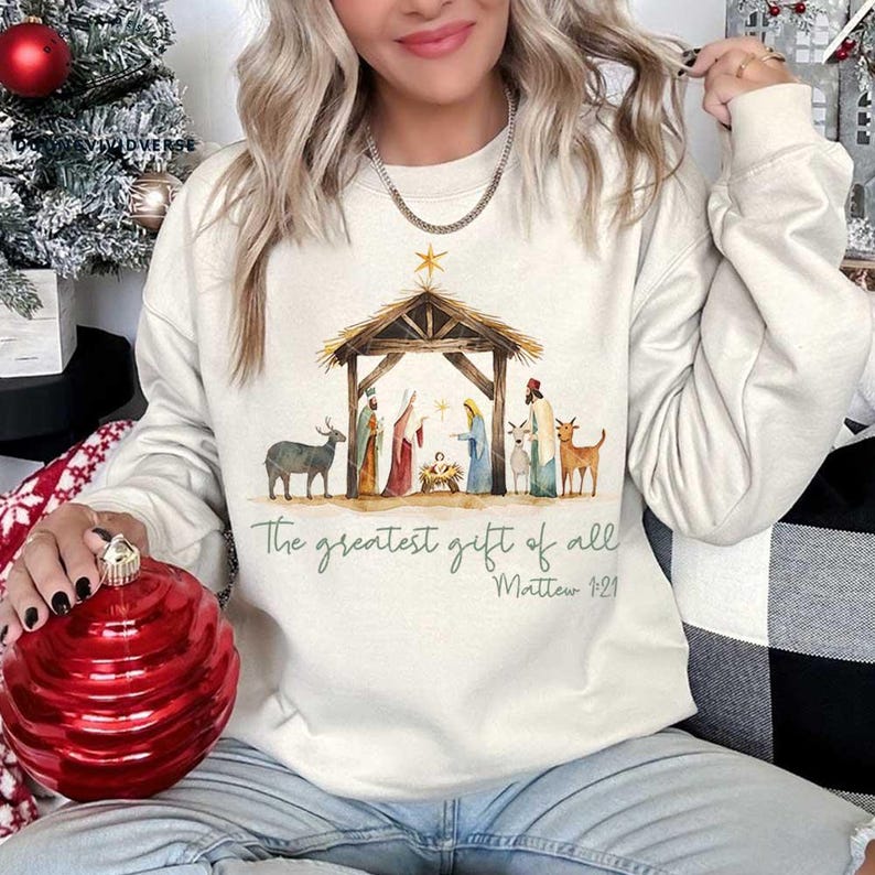 May include: A white sweatshirt with a watercolor illustration of the nativity scene. The text "The greatest gift of all" and "Matthew 1:21" is printed below the image.