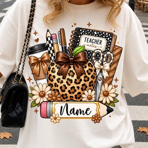 May include: A cream-colored t-shirt featuring a graphic print of a leopard-print apple with a brown bow, a notebook labeled Teacher Notes, a coffee cup, scissors, and a pencil with the word Name written in black cursive script.