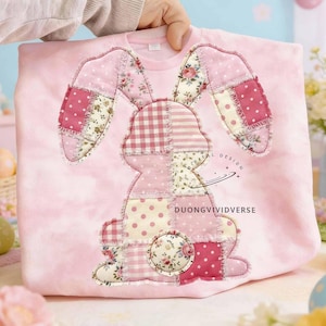 May include: A light pink sweatshirt featuring a patchwork bunny design. The bunny is made of various pink fabrics with floral and polka dot patterns. The text "DUONGVIVIDVERSE" is visible. A hand is holding the sweatshirt.