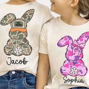 May include: Two white t-shirts with a camouflage bunny design. The bunny on the left is wearing a hat that says "Egg Hunter". The bunny on the right has a pink bow. The shirts are personalized with the names "Jacob" and "Sophia".