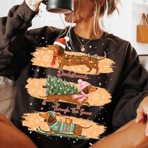 May include: A dark brown long-sleeve sweatshirt featuring three dachshunds in winter attire. The top dog wears a Santa hat and lights, the middle carries a Christmas tree, and the bottom wears a sweater and skis. Text reads Dachshund Through The Snow.
