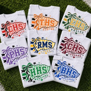 Pode incluir: Uma coleção de oito camisetas brancas, cada uma apresentando um gráfico de flâmula colorido com um laço grande, iniciais da escola e nomes de mascotes, incluindo Vikings, Tigers, Eagles, Lions, Cougars, Panthers, Warriors e Bulldogs em vários padrões xadrez.