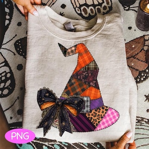 May include: A light gray sweatshirt featuring a colorful patchwork witch hat design with a black bow. The hat incorporates plaid, floral, and leopard print patterns in orange, purple, and pink. A black bow with gold stars and orange accents is at the base.