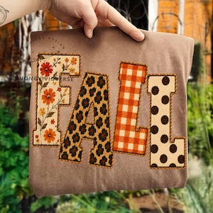 May include: A brown t-shirt featuring the word FALL in large block letters. Each letter has a distinct pattern: the F has orange and yellow flowers, the A has a dark leopard print, the L has an orange gingham check, and the L has brown polka dots.