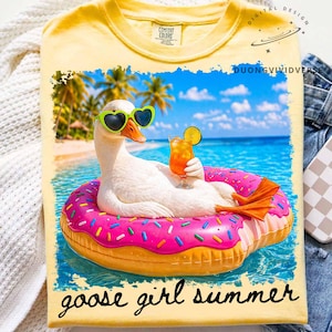 May include: Yellow t-shirt featuring a goose wearing heart-shaped sunglasses and floating on a pink donut-shaped floatie in the ocean. The goose is holding a cocktail. The words "goose girl summer" are printed at the bottom.