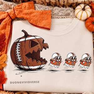 Puede incluir: Una sudadera color crema con un gráfico de una calabaza de Halloween con forma de balón de fútbol americano persiguiendo a tres pequeños fantasmas con cascos y camisetas de fútbol naranja y blanco. El texto DUONGVIVIDVERSE aparece abajo.