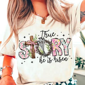 May include: White t-shirt with a floral design that says "True Story" with a cross, nails, and a crown of thorns. The text below says "He is risen".