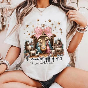 May include: White t-shirt with a pink bow and a nativity scene with the text "The greatest gift of all Matthew 1:21".