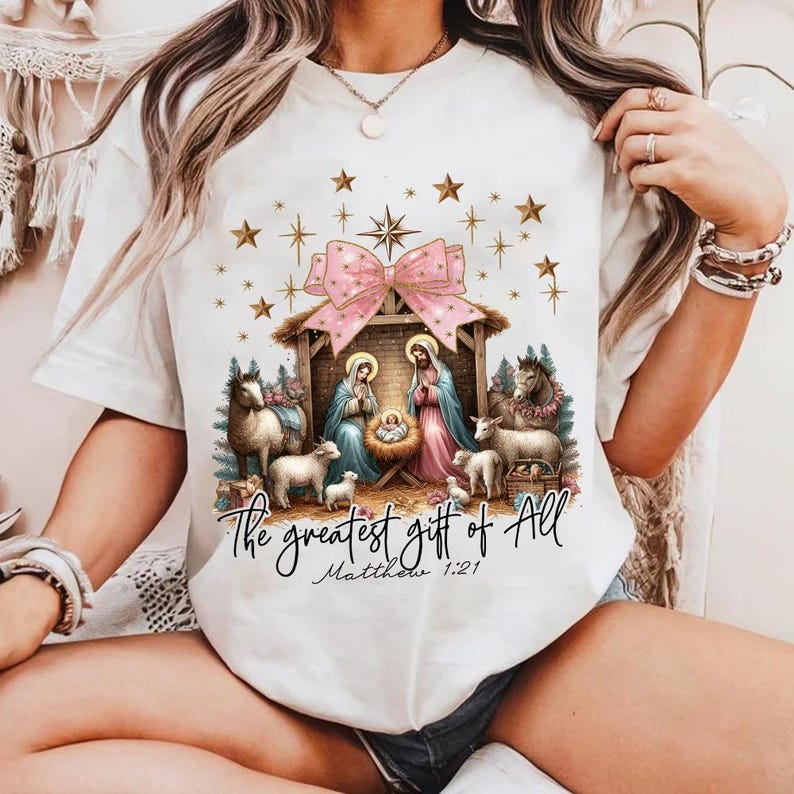 May include: White t-shirt with a pink bow and a nativity scene with the text "The greatest gift of all Matthew 1:21".