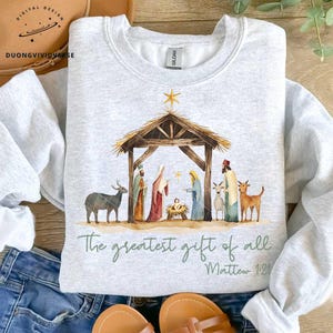 May include: A gray sweatshirt with a watercolor illustration of the nativity scene. The illustration includes a manger, a star, a donkey, a sheep, and figures of Mary, Joseph, and the baby Jesus. The text "The greatest gift of all" and "Matthew 1:21" is printed below the illustration.