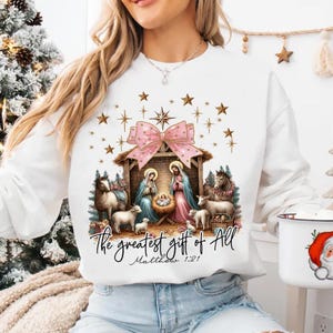 May include: White Christmas sweatshirt with a pink bow and a nativity scene. The text reads "The greatest gift of all Matthew 1:21".