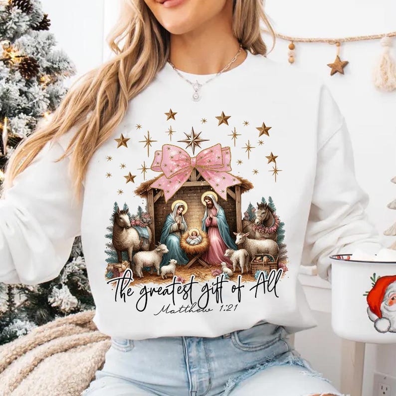 May include: White Christmas sweatshirt with a pink bow and a nativity scene. The text reads "The greatest gift of all Matthew 1:21".