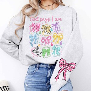 May include: A gray sweatshirt with the text "God says I am" printed on the front. The text is surrounded by colorful bows with different bible verses and words like "unique", "loved", "precious", and "strong".