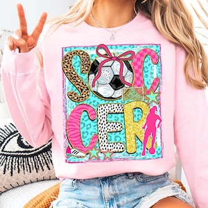 Op de afbeelding: Zachtroze sweatshirt met een kleurrijk voetbalthema. Het woord "SOCCER" is geschreven met verschillende patronen en een voetbal. De afbeelding bevat een voetbal, een doel, sterren en een silhouet van een voetballer.