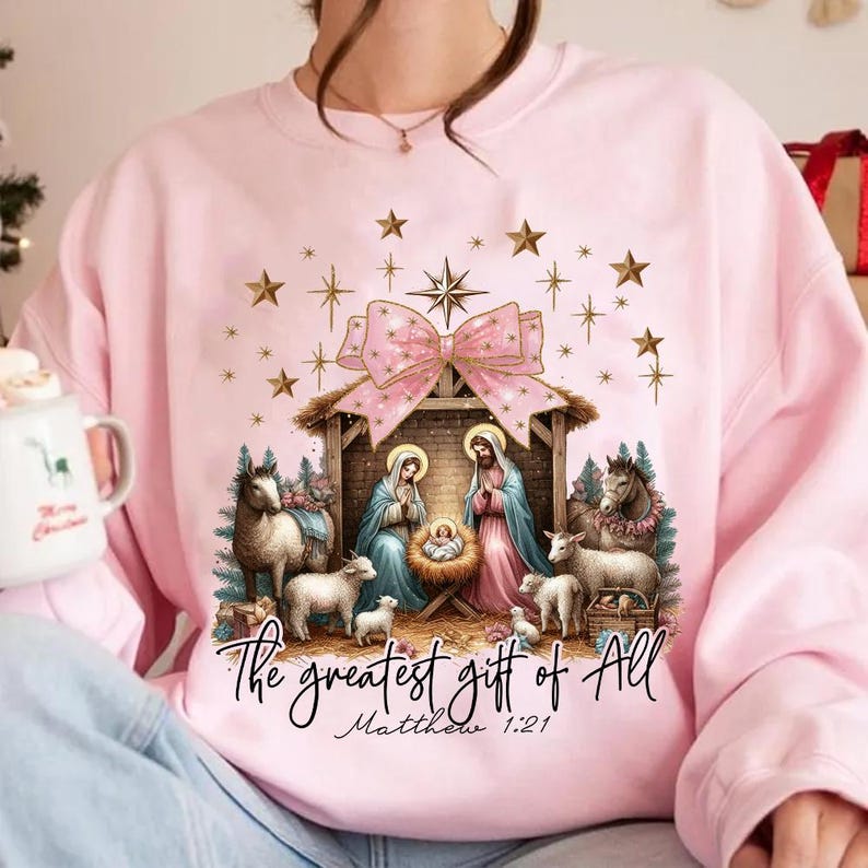May include: Pink sweatshirt with a Christmas nativity scene design. The scene features Mary, Joseph, and baby Jesus in a manger with a pink bow and gold stars. The text "The greatest gift of all Matthew 1:21" is printed below the scene.