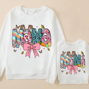 May include: Two white sweatshirts with long sleeves. The larger sweatshirt says "Mama" in colorful, patterned letters with a pink bow. The smaller sweatshirt says "Mini" in a similar style. Both have Easter-themed graphics.