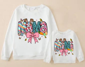 Puede incluir: Dos sudaderas blancas de manga larga. La sudadera más grande dice "Mama" en letras coloridas y estampadas con un lazo rosa. La sudadera más pequeña dice "Mini" en un estilo similar. Ambas tienen gráficos temáticos de Pascua.