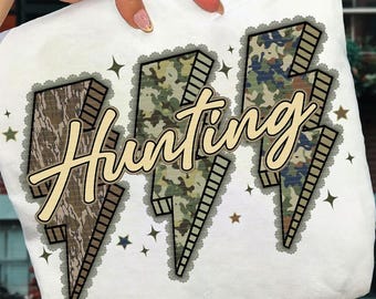 May include: White t-shirt with the word "Hunting" in a beige script font. The word is superimposed on three lightning bolt shapes with camouflage patterns and a decorative border. Small star shapes are scattered around the design.