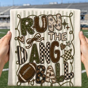 May include: A cream-colored fabric featuring the text RUN THE DANG BALL in large, patterned letters. The design includes football-themed graphics like helmets, a football, and stars, all connected by a thin, winding rope-like line pattern.