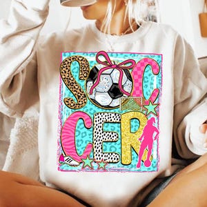 Op de afbeelding: Witte sweatshirt met een kleurrijke "SOCCER"-afbeelding. Het ontwerp bevat een voetbal met een roze strik, een net, sterren en een silhouet van een voetballer. De letters hebben verschillende patronen en kleuren.