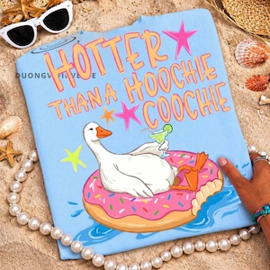 Op de afbeelding: Lichtblauwe strandlaken met de tekst "Hotter Than a Hoochie Coochie" in oranje en roze. Een cartoon gans drijft op een roze donut, met een cocktail. Zonnebril en schelpen op de achtergrond.