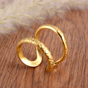 Peut inclure: Une bague dorée avec une surface texturée. La bague présente un design unique, avec une bande incurvée qui se connecte à une seconde bande. La bague est présentée sur une surface en bois.