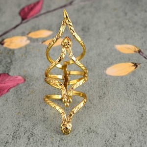 May include: A gold-colored ring with an intricate design. The ring features a series of interconnected loops and curves, with a small, orange-colored stone at the center. The ring's design is detailed and ornate, with a textured surface.