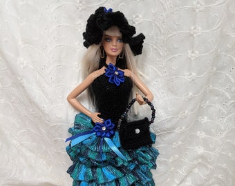 May include: A doll wearing a black halter dress with a layered teal and blue skirt. The doll has a black crocheted hat and a matching purse. Blue ribbon and flower accents adorn the dress.
