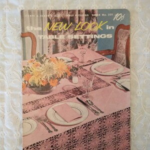 May include: Vintage magazine cover titled "The New Look in Table Settings." The cover features a pink tablecloth with a crocheted border, set with plates, silverware, and glassware. A floral arrangement and the text "10¢" are also visible.