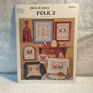 May include: A book cover titled "plain & fancy FOLK 2" by Jeremiah Junction. The cover features framed cross-stitch samplers and decorative items. The book is labeled "Book 9" and includes the text "COUNTED CROSS STITCH".