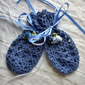 May include: A pair of blue crocheted mittens tied with blue and white ribbons. The mittens are decorated with small fabric roses and pearls. The openwork design of the mittens is visible.