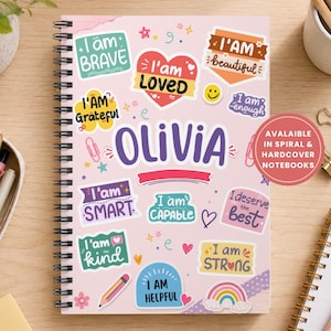 May include: A spiral-bound notebook with a light pink cover featuring the name OLIVIA in large purple letters. The cover is decorated with colorful stickers containing positive affirmations like I am BRAVE, I am LOVED, and I am STRONG.