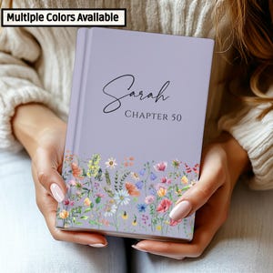 Puede incluir: Un diario lavanda con el nombre "Sarah" y "CHAPTER 50" impresos en la portada. La parte inferior del diario presenta un diseño floral con flores coloridas. El texto "Multiple Colors Available" está en la esquina superior izquierda.