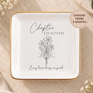 May include: A square white ceramic trinket dish with a gold-colored rim. The center features a black line drawing of a daffodil bouquet, the text "Chapter EIGHTEEN", the name "EMMA" inside a horizontal line frame, and "Every chapter brings new growth".