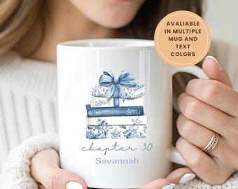 May include: White ceramic mug featuring a stack of books with a blue bow and floral designs. The mug has the text "chapter 30" and the name "Savannah" in blue. The mug is available in multiple colors.