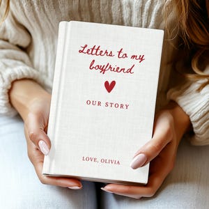 May include: A cream-coloured book with the title "Letters to my boyfriend" in red script. A red heart is centered below the title, with "OUR STORY" beneath it. The book is personalised with "LOVE, OLIVIA" at the bottom.