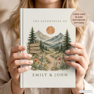 May include: A white hardback notebook titled The Adventures of Emily & John, featuring a watercolour illustration of a mountain landscape, pine trees, a dirt path, and wildflowers. A wooden signpost reads Places, People, Memories.